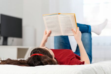 A young Caucasian woman is lying on the bed on her back, reading a book. White interior of the room on the background. Concept of education and world book day.の写真素材