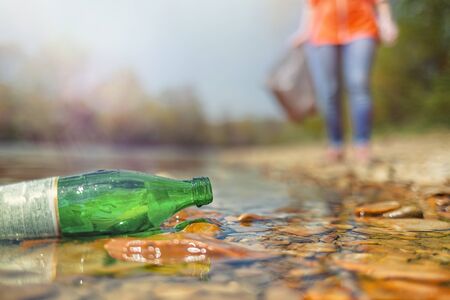 On the banks of the river is a glass bottle thrown by someone. In the background is a volunteer. The concept of environmental protection and sustainability. Light.の写真素材