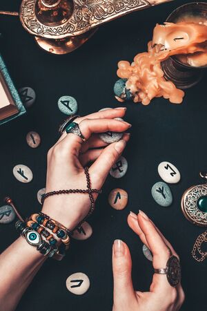 Astrology and esotericism. The witch is guessing on Runestones. Hands close up. On a black background lie fortune-telling runes, precious amulets, a copper lamp and a candle. Vertical.の写真素材