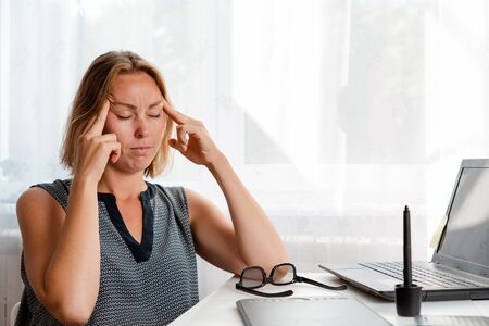 A woman sitting at a work Desk, rubbing her temples from a headache. White window in the background....
