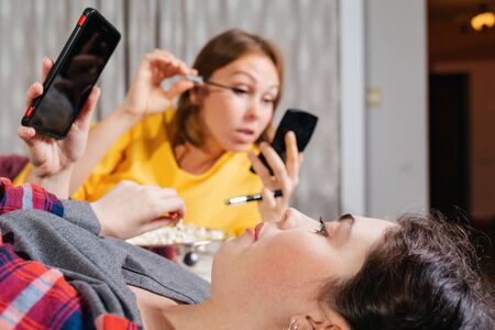 Two young women are lying on the bed, eating popcorn, using smarfton, and decorative cosmetics are...