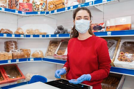 A portrait of woman in rubber gloves and a medical mask puts money in the cash register. In the...
