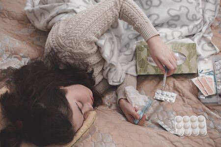 a sick girl in a sweater lying on the bed checks the temperature with a thermometer on the background of pills scattered on the blanket.の写真素材