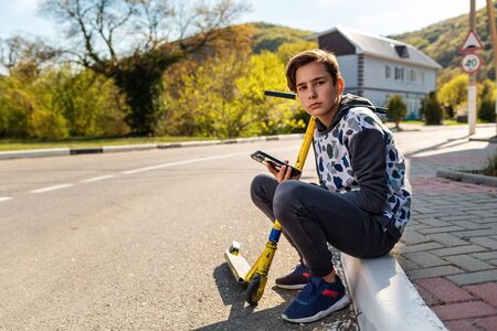 A brooding teenage boy sits on the sidewalk with a scooter and a mobile phone in his hand. In the background, an empty street, a Sunny day. The concept of modern technologies, activity and young people.の写真素材