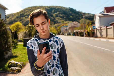 A Caucasian teenage boy looks thoughtfully at the smartphone in his hand. In the background, a street, a Sunny day. Concept of communications and modern technologies.の写真素材