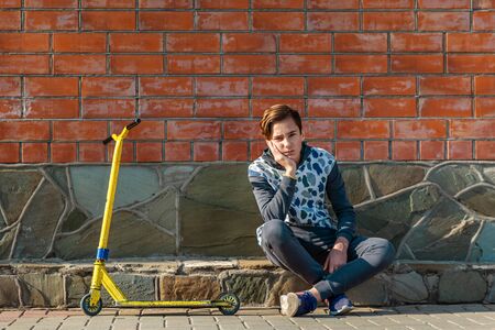 A teenage boy sits on the sidewalk next to a scooter, and frowns leaning on his hand. Brick wall in...