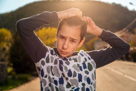 A teenage boy ties his hair in a ponytail with an embarrassed look. Emotion. Street in the background. The concept of hairstyles and self-care.の写真素材