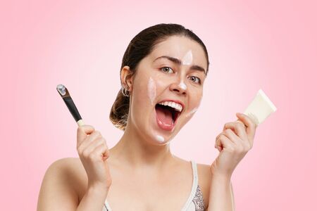 Portrait of a young beautiful woman with a happy smile, holding a tube with a mask and a brush in...