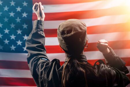 Veterans Day, Memorial Day, Independence Day. A female soldier rejoices, raising her hands to the top, against the background of the American flag. Rear view. The concept of the American national holidays and patriotism.の写真素材