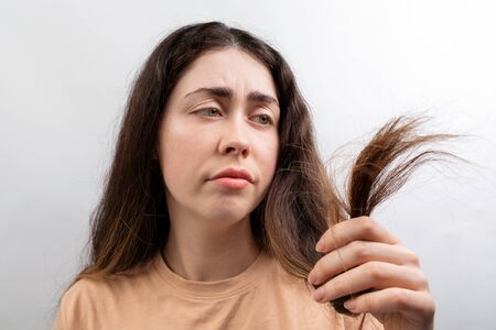 A. young Caucasian woman in a beige t-shirt looks distressed at the dry ends of her long hair. White...