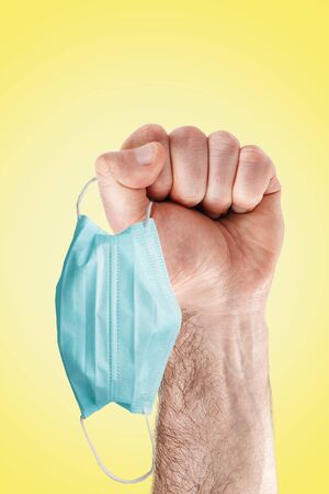 Covid-19. A man's hand holds a medical mask in his fist. A symbol of the fight against the pandemic. Yellow background. Concept of quarantine and virus protection.の写真素材