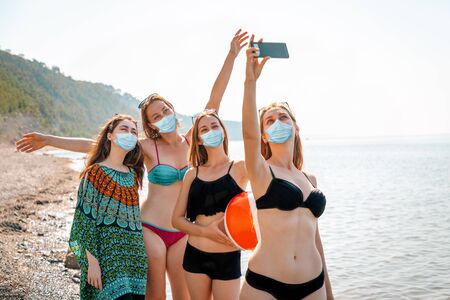 Covid-19. A group of young women in bathing suits and medical masks take selfies on a smartphone. The sea in the background. The concept of home away from home during a pandemic, and protect against infection.の写真素材