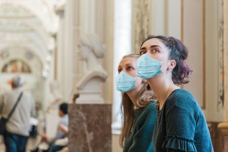 Coronavirus. Two women in a medical mask, admiring the masterpieces in the Museum, being at a distance from other people. The concept of protection from the virus while visiting public places.の写真素材