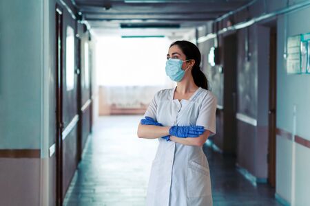 Coronavirus.Doctor a woman in a lab coat, medical mask and gloves poses in a hospital corridor with her arms crossed over her chest.The concept of hero doctors and the stress of overloading at work during the pandemic.の写真素材