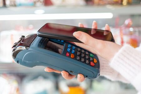 A woman's hands is holding a payment terminal and paying for a purchase using a smartphone. Copy space and side view. The concept of NFC, business and banking transactions.の写真素材