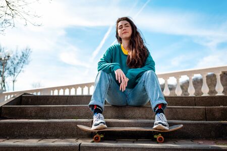Cute Caucasian teen girl sitting on the steps and leaning her feet on a skateboard. In the background, the blue sky and the Boulevard. Bottom view. Concept of sports lifestyle and street culture.の写真素材