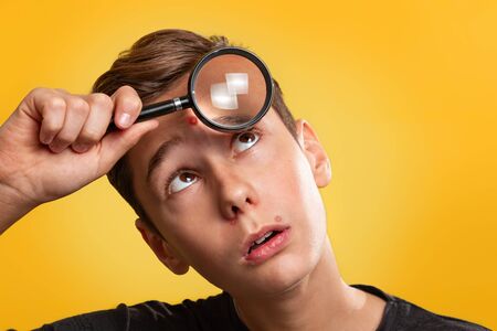Cosmetology, Dermatology and acne. Teenage boy in a black t-shirt with pimples on his face, holding a magnifying glass to his forehead. Yellow background. Close up.の写真素材