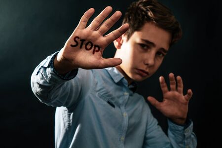 The concept of domestic violence. Sad teenager puts his hands in front of him defending himself from the threat of attack. Black background. Text STOP .の写真素材