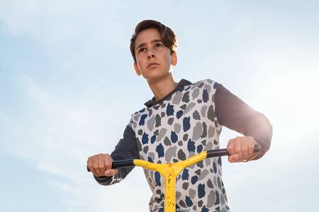 A teenage boy with a serious face rides a scooter. Blue sky in the background. Bottom view. Concept of sports recreation and youth activity.の写真素材