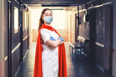 Doctor wearing medical facemask, gloves and superhero cape during coronavirus outbreak. Virus and illness protection, quarantine. Corridor of hospirtal on the background. COVID-2019. Concept of Super hero power for medicine.の写真素材