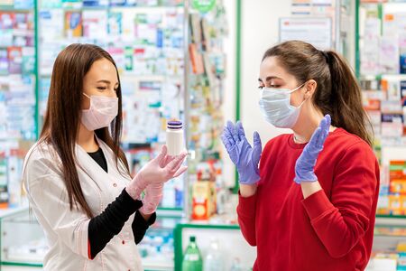 The pharmacist shows the good medicine to the buyer.Two women in medical masks and gloves on the background of a window with pharmacy products. Emotions of surprise. The concept of drug purchase in a pharmacy.の写真素材