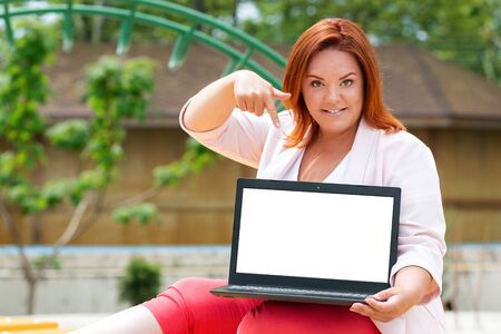 Freelance. Portrait of caucasian smiling woman holds a laptop and points at the screen with her finger. Plus size model. Outdoor. Mock up. Concept of remote work and online businessの写真素材
