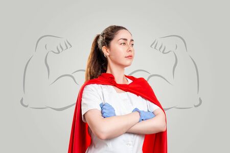 A doctor in surgical gloves and a red superhero Cape, posing against a background of painted strong hands on a gray background.の写真素材