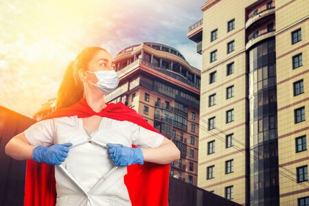 A doctor wearing medical gloves, a surgical mask, and a red superhero Cape rips the medical gown on his chest. Modern buildings in the background. The concept of the Power of a super hero for medicine.の写真素材
