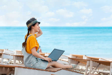 Freelance. A young woman sits relaxed on a sunbed with a laptop on her lap and looks out at the sea. Summer holiday. Copy space.の写真素材