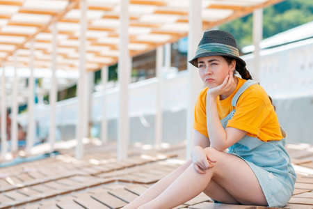 Emotion of sadness and discontent. Portrait of young woman in a straw hat leaned on her hand in discontent, sitting in the shade on the sun beds.の写真素材