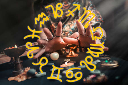 Astrology and esotericism. The witch at the candle and rune stones. Sparks of fire in the air. Dark background. Yellow zodiac horoscope at the foreground.の写真素材