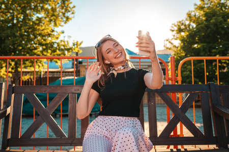 A happy Woman sits on a bench in a Park and communicates via video link on a smartphone. Summer. The concept of social dependence and telecommunications.の写真素材