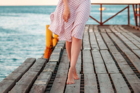 Summer vacation. A slim woman in a dress is walking on tiptoe along the pier by the sea. Legs close up.の写真素材