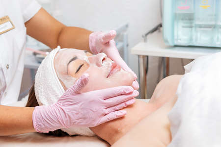 A beautician wearing rubber gloves applies a woven mask to a woman's face. Close-up of a satisfied customer's face. Concept of cosmetology and treatment.の写真素材
