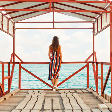 Summer holiday. A woman in a striped dress poses on the pier. Rear view. The sea is in the background.の写真素材