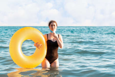 Social distance and virus protection. A woman in a bathing suit removes a medical mask and poses with an inflatable circle while standing in the sea. In the background, the sea and sky, the horizon.の写真素材