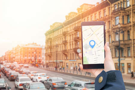 A woman's hand in a jacket holds a smartphone with an online map app. In the background is a city...