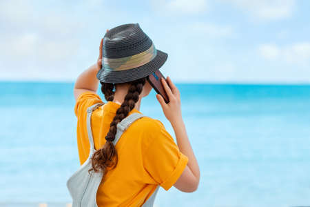A woman in a straw hat call at the mobile phone. Rear view. Sea and sky in the background. Copy space. Concept of vacations and communications.の写真素材