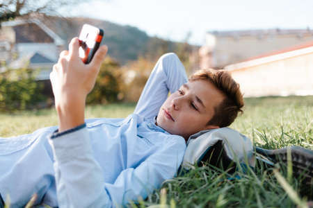 Mobile phone and Internet. A teenage boy lies in the Park on the grass and watches a video on a smartphone.の写真素材
