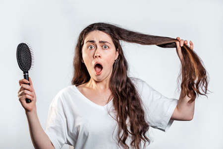 A frightened woman with long dark hair, screaming, holding a comb and a lock of her long hair. White background. Concept of hair loss, baldness and hair care.の写真素材