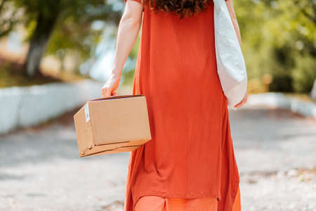 A woman in a red dress is walking down the street with a parcel in her hand. Rear view. The concept of delivery of goods.の写真素材