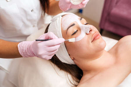 The cosmetologist in medical gloves holds a bowl with cosmetics, applying a brush mask on the client's face. Concept of professional cosmetology.の写真素材