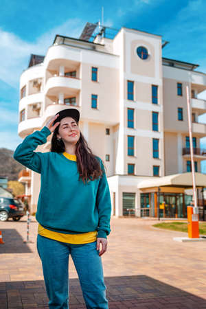 A young Caucasian woman in casual clothes holds her cap with a smile. In the background is a new...