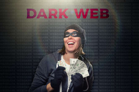 The concept of cybercrime and illegal outlets. Portrait of a woman in a black hat, gloves and mask, who enthusiastically holds a wad of money. Dark background with backlight and the words dark web.の素材 [FY310154462894]