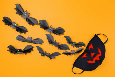 Reusable anti-virus mask with print of jack lantern surrounded by paper bats. Orange background. Copy space. Flat lay. The concept of protection from the virus during Halloween.の写真素材