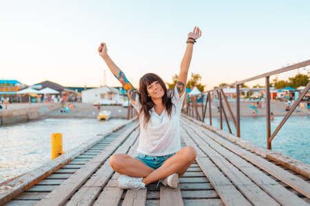 A pretty woman with tattoos sits cross-legged on a wooden pier by the sea, her hands raised in joy....
