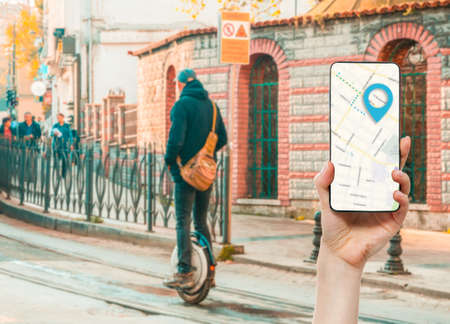A hand holds a smartphone with an online maps app. In the background, a man rides down the street on...