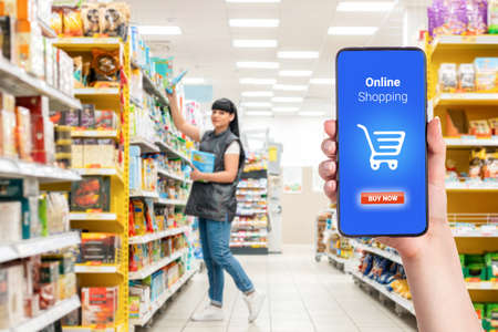 A person's hand holds a mobile phone, with a shopping basket on the screen. In the background, a...