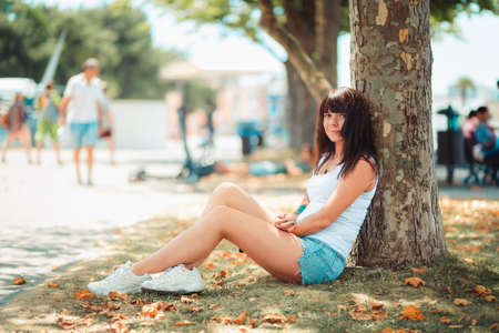 Summertime. A smiling woman is sitting relaxed leaning against a tree trunk. Recreation in the Park.の写真素材
