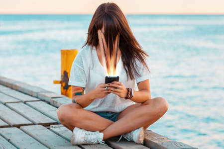A woman sits on a pier by the sea and watches social media on her smartphone. Face smudged, sucked into the phone. The concept of online addiction.の写真素材